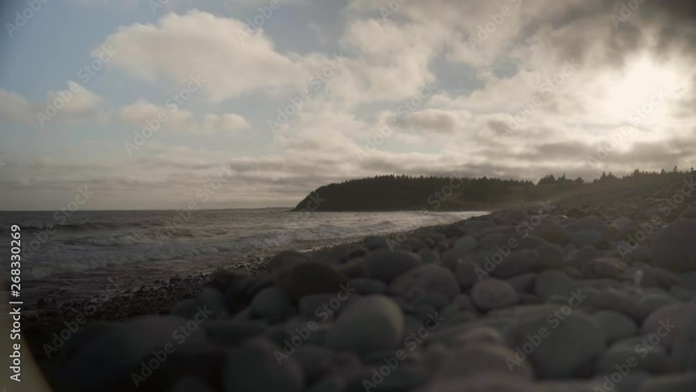 Ground shot of some waves hitting beach with some stones on the ground, trees and a road on the background near sunset in Halifax, Nova Scotia, Canada. Ground shot of some waves hitting beach with some stones on the ground, trees and a road on the background near sunset in Halifax, Nova Scotia, Canada.