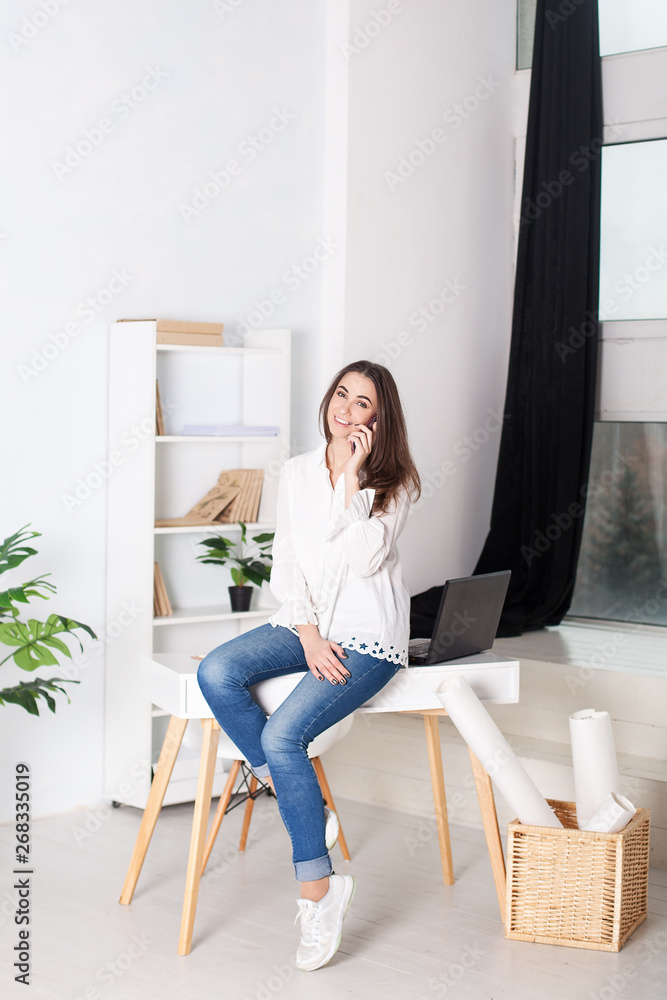 Business vumen. Young girl working in the office. The girl calls on the phone and sits on the table. Calling customers of the company. Positive young manager working on business project in office. Business vumen. Young girl working in the office. The girl calls on the phone and sits on the table. Calling customers of the company. Positive young manager working on business project in office.