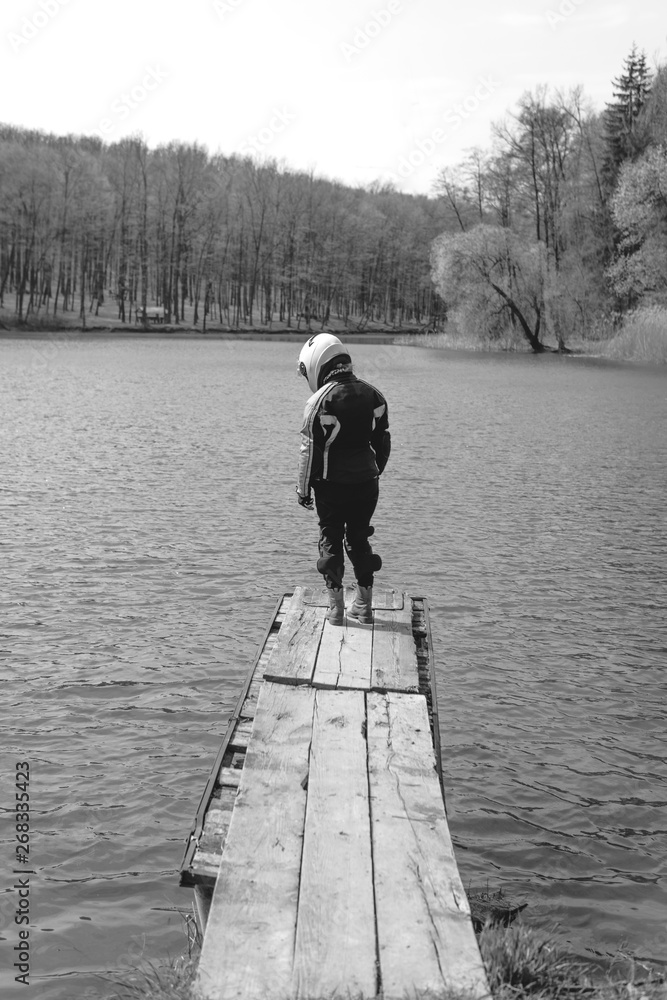 A sad girl is standing alone on the pier by the lake. Forest background. wearing in motorcycle outfit protection equipment. Emotions of a depresion of loneliness, problems in social adaptation