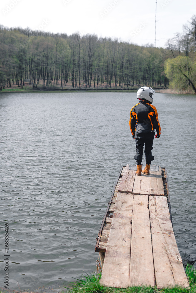 A sad girl is standing alone on the pier by the lake. Forest background. wearing in motorcycle outfit protection equipment. Emotions of a depresion of loneliness, problems in social adaptation