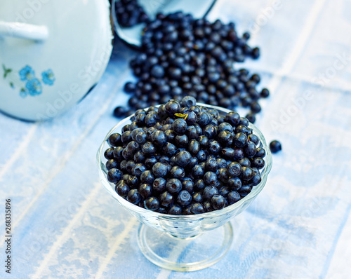 A glass vase full of fresh forest blueberry on the background of berries pouring out of the milk can, everything on blue with white linen rustic textile, close-up, copy space