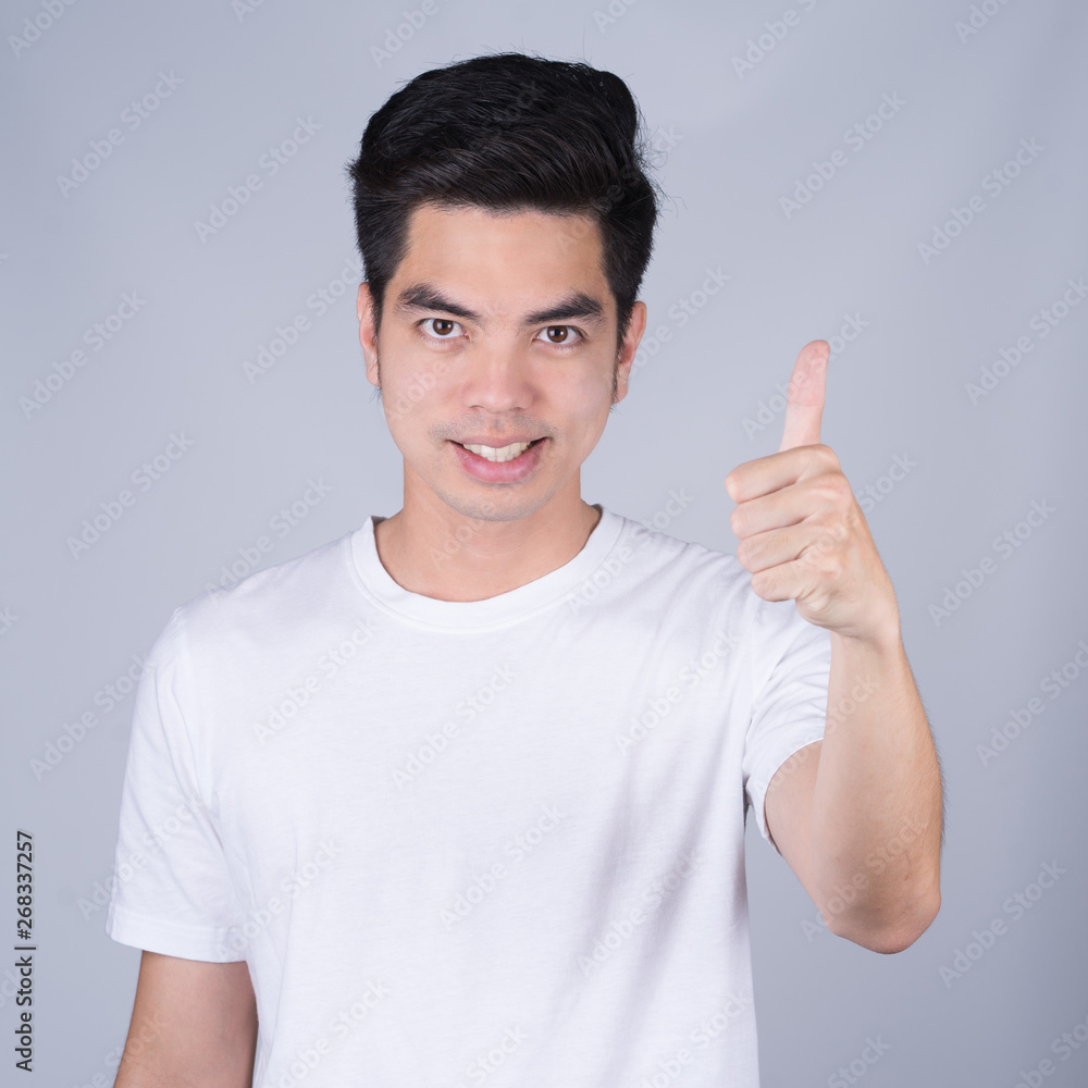 Portrait handsome young asian man wearing a white T-shirt showing thumbs up sign isolated on grey background. Asian man people. business success concept. Portrait handsome young asian man wearing a white T-shirt showing thumbs up sign isolated on grey background. Asian man people. business success concept.