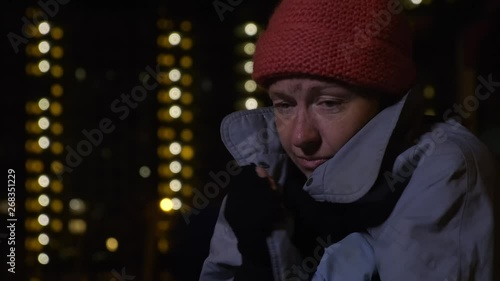Closeup of frozen homeless man wraps himself in coat against background of night city.