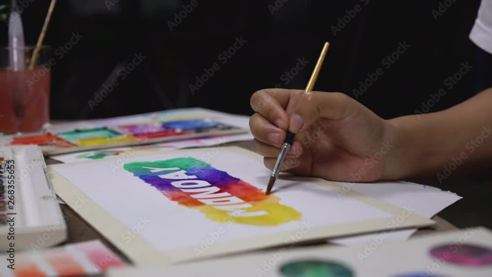 Low angle selective focus view of an artist at work painting a watercolour design on canvas - Hand view of a young creative hand painting colour to an artwork in a dark art studio Low angle selective focus view of an artist at work painting a watercolour design on canvas - Hand view of a young creative hand painting colour to an artwork in a dark art studio