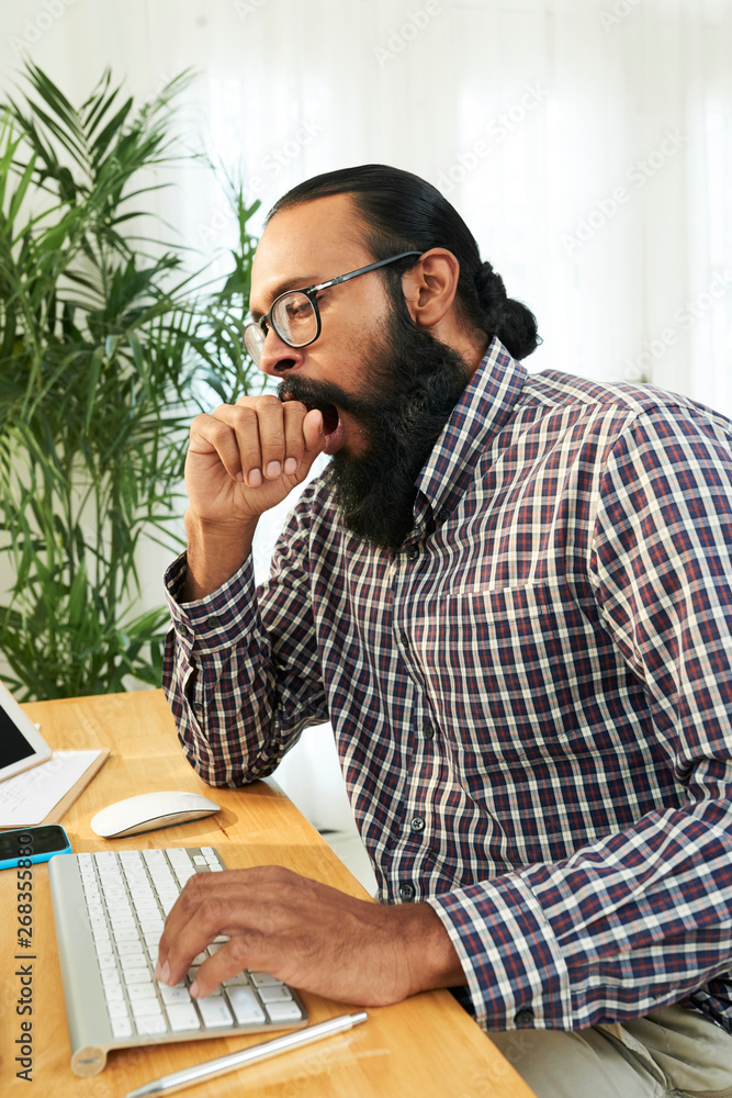Fototapeta premium Bearded young manager yawning while typing on computer keyboard at office desk