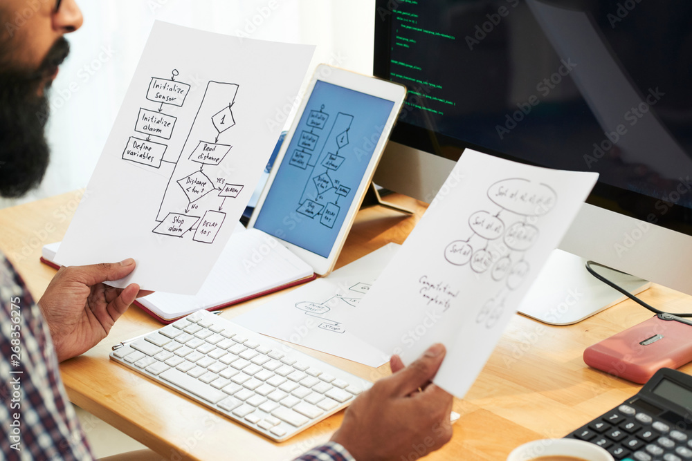 Close-up of bearded manager sitting at his workplace and examining two business strategies on papers in his hands at office Close-up of bearded manager sitting at his workplace and examining two business strategies on papers in his hands at office