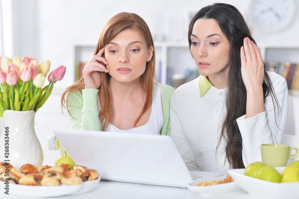 Portrait of two young women looking at laptop while sitting at kitchen table with cookies