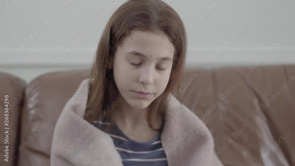 Close up portrait of teenage girl wrapped in a blanket blowing her nose in a napkin sitting on the leather sofa at home. The girl feels bad, she is ill and has a fever. Concept of healthcare Close up portrait of teenage girl wrapped in a blanket blowing her nose in a napkin sitting on the leather sofa at home. The girl feels bad, she is ill and has a fever. Concept of healthcare