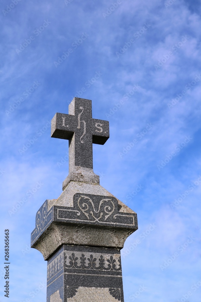 Looking up at an old stone grave monument with blue sky and wispy clouds in behind with i.h.s. representing Jesus Christ