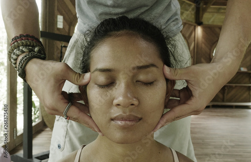 close up face portrait of young gorgeous and relaxed Asian Indonesian woman receiving traditional facial Thai massage with male hands working her head