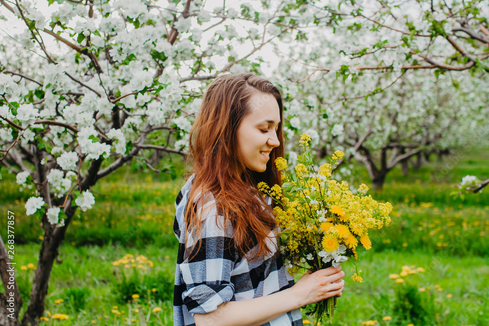 A girl with a yellow spring bouquet of wildflowers in her hands stands sideways in a blooming Apple orchard looking at the flowers and smiling A girl with a yellow spring bouquet of wildflowers in her hands stands sideways in a blooming Apple orchard looking at the flowers and smiling