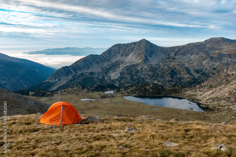 Orange tent in mountains with grass, lake and mountains in background during sunrise