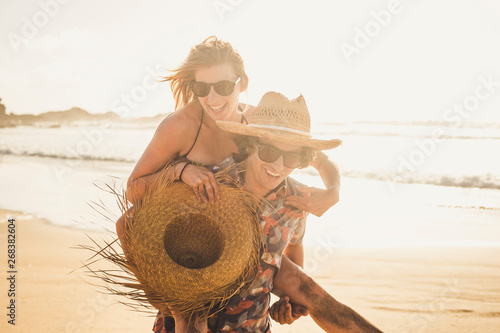 Young cheerful people having fun in couple with man carry beautiful woman - laughing together and enjoying the sunny day of holiday vacation at the beach - romance and love concept
