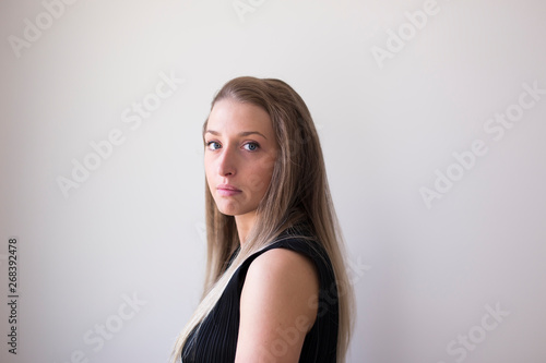 Horizontal closeup of gorgeous serious-looking blonde young woman in sleeveless black top turning back and staring against plain cream background