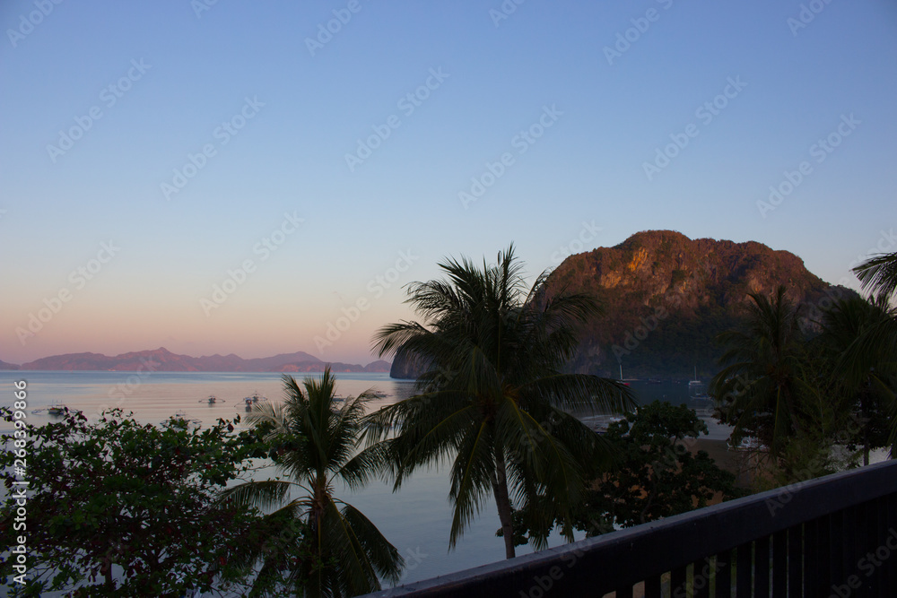 Beautiful view of tropical morning bay from balcony. Morning on Philippines island with trees and boats. Sunrise on Palawan, Philippines. Tropical isles in morning dusk with trees on foreground. Asian Beautiful view of tropical morning bay from balcony. Morning on Philippines island with trees and boats. Sunrise on Palawan, Philippines. Tropical isles in morning dusk with trees on foreground. Asian
