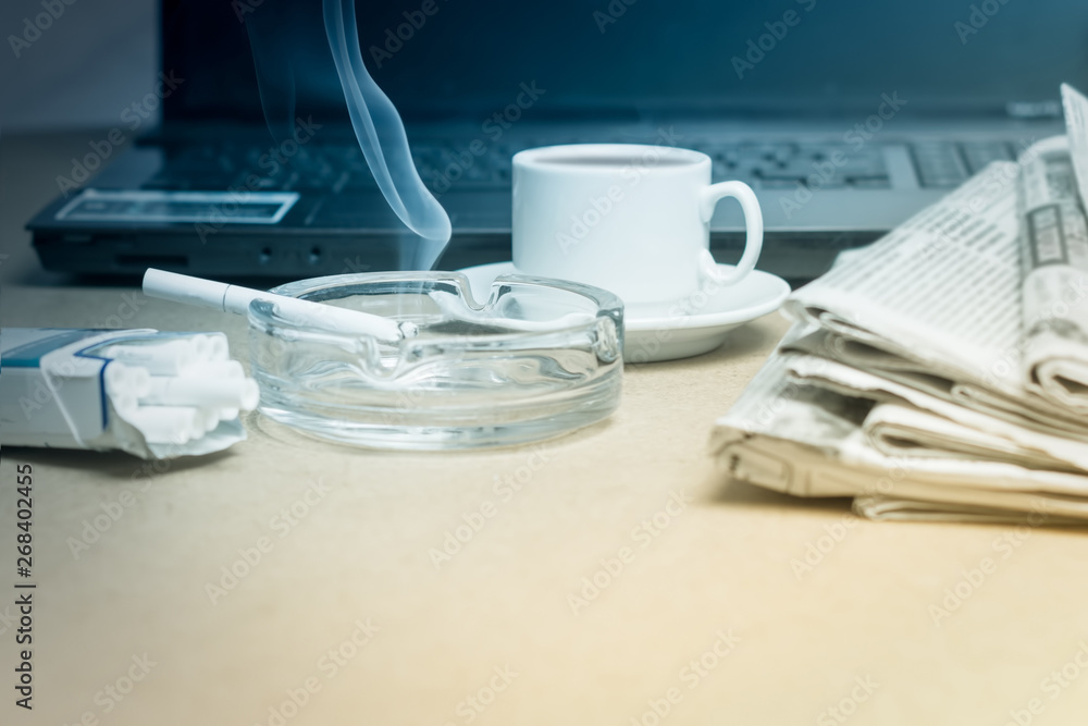 The concept of news. A folded stack of Newspapers on the laptop, a Cup of coffee, a Smoking cigarette in the ashtray. Office worker's Desk. The concept of news. A folded stack of Newspapers on the laptop, a Cup of coffee, a Smoking cigarette in the ashtray. Office worker's Desk.