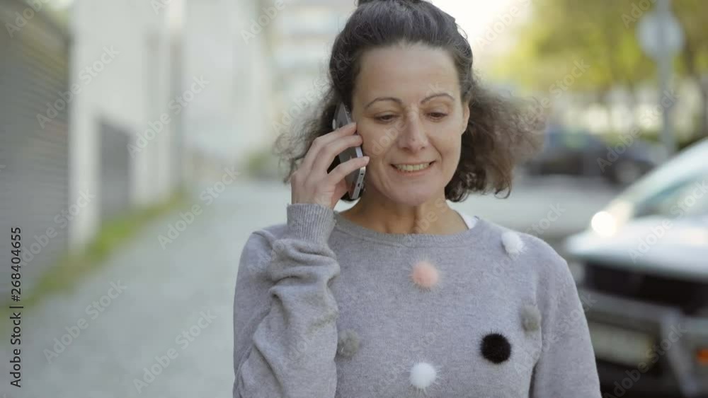 Relaxed woman talking on phone while strolling on street. Smiling mature lady talking with friend through smartphone. Communication and technology concept Relaxed woman talking on phone while strolling on street. Smiling mature lady talking with friend through smartphone. Communication and technology concept