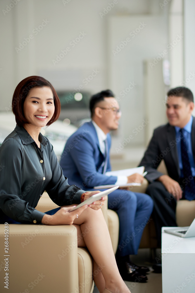 Asian young businesswoman sitting on sofa with tablet pc and smiling at camera while has a business meeting with her colleagues in car showroom Asian young businesswoman sitting on sofa with tablet pc and smiling at camera while has a business meeting with her colleagues in car showroom