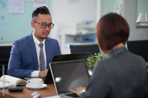 Asian serious businessman in eyeglasses sitting at office desk and typing on laptop computer with businesswoman working opposite him