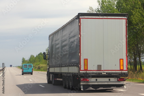 Transportation logistics - truck with gray semi-trailer moving on asphalt road in summer day against blue sky forest, rear side view
