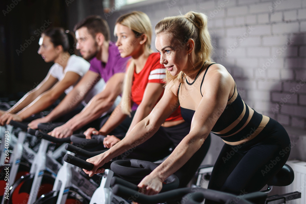Group of smiling friends at gym exercising on stationary bike. Happy cheerful athletes training on exercise bike. Young men and woman working out at a class in the gym. Group of smiling friends at gym exercising on stationary bike. Happy cheerful athletes training on exercise bike. Young men and woman working out at a class in the gym.