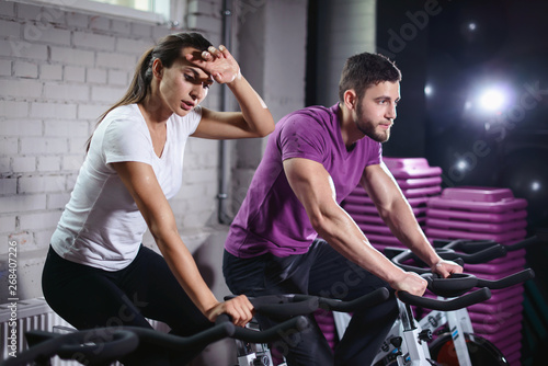 Attractive woman and man biking in the gym, exercising legs doing cardio workout cycling bikes. Couple in a spinning class wearing sportswear. Fitness healthy lifestyle concept
