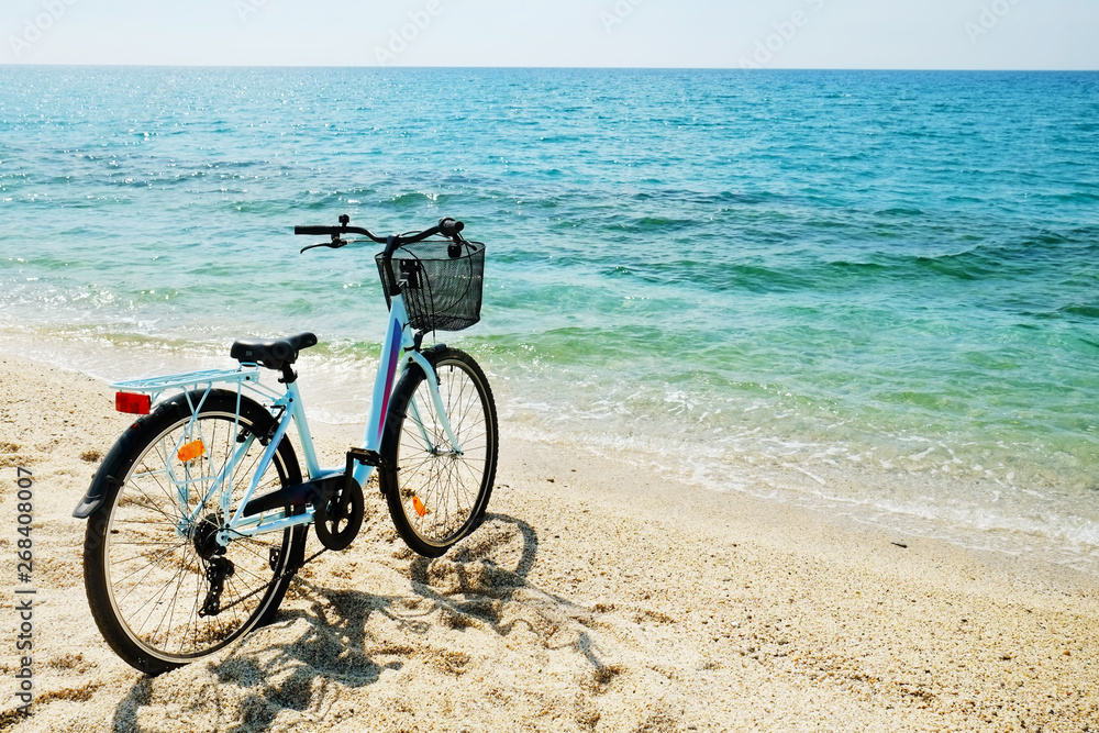 Fototapeta premium Feminine bicycle of comfort class with empty basket on the sandy beach of mediterranean sea. Blue cruiser bike on sunny day at sea shore with a lot of copy space for text.
