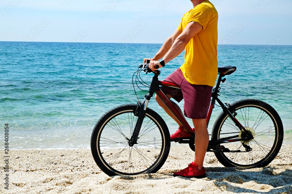 Young man with fit body riding the mtb mountain bike on sandy beach with beautiful azure water sea view. Muscle male wearing bright yellow t-shirt cycling on ocean shore. Close up, copy space. Young man with fit body riding the mtb mountain bike on sandy beach with beautiful azure water sea view. Muscle male wearing bright yellow t-shirt cycling on ocean shore. Close up, copy space.