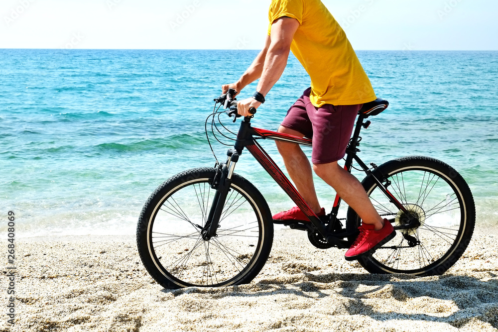 Fototapeta premium Young man with fit body riding the mtb mountain bike on sandy beach with beautiful azure water sea view. Muscle male wearing bright yellow t-shirt cycling on ocean shore. Close up, copy space.