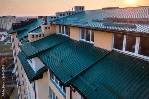 Aerial view of attic annex room exterior with plastic windows, roof and walls covered with green metal siding planks, new gutter system on top of high multi-storey apartment building.