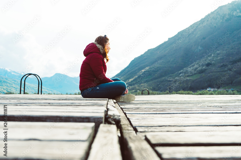 A young girl sits on the edge of a wooden pier and looks into the distance surrounded by mountains in Plav lake A young girl sits on the edge of a wooden pier and looks into the distance surrounded by mountains in Plav lake