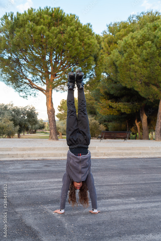 Obraz premium Young handsome flexible male in sportswear with roller skates doing handstand on road in beautiful city park