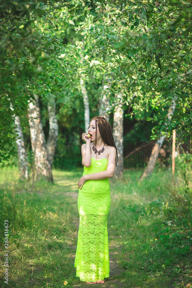 Barefoot girl with dreadlocks in a long green dress with a green apple in hand in the garden among the apple trees Barefoot girl with dreadlocks in a long green dress with a green apple in hand in the garden among the apple trees