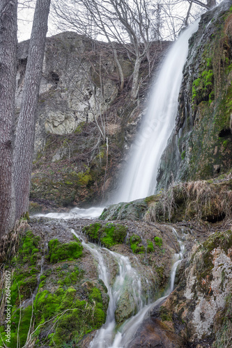 Close up, long exposure view of a sunlit waterfall with soft streams of water falling down red rocks covered by green moss
