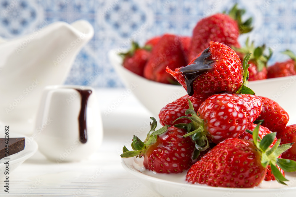 Close up. White saucer with a slide of juicy strawberries. A bitten berry lies on top, with a drop of chocolate flowing from it. Capacity with dripping chocolate on the background. Close up. White saucer with a slide of juicy strawberries. A bitten berry lies on top, with a drop of chocolate flowing from it. Capacity with dripping chocolate on the background.
