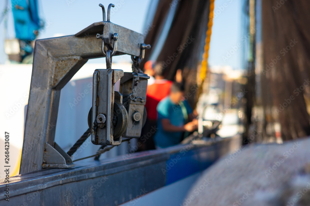 A winch on fishing boat in aftermath. In the background, in the afternoon, the fishermen of Torrevieja prepare the black fishing net on their fishing boat for their next fishing trip at night. A winch on fishing boat in aftermath. In the background, in the afternoon, the fishermen of Torrevieja prepare the black fishing net on their fishing boat for their next fishing trip at night.