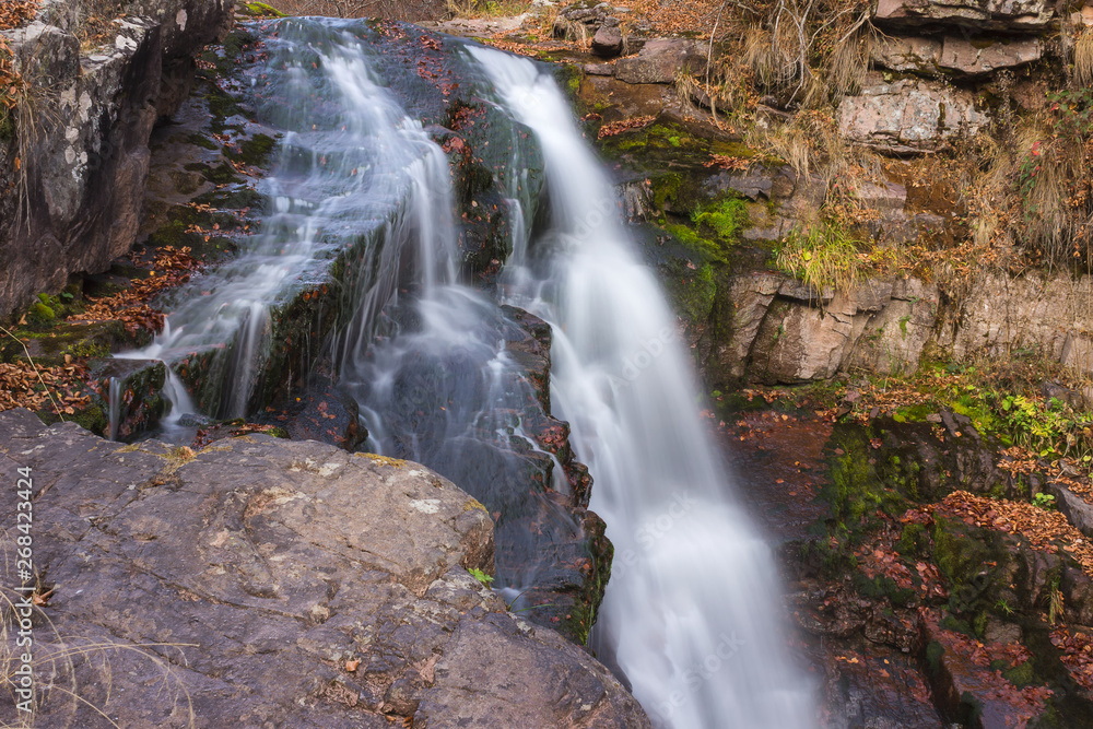 Fototapeta premium Gorgeous mountain creek waterfall cascading down the rocky cliff and colorful autumn red leaves, close-up