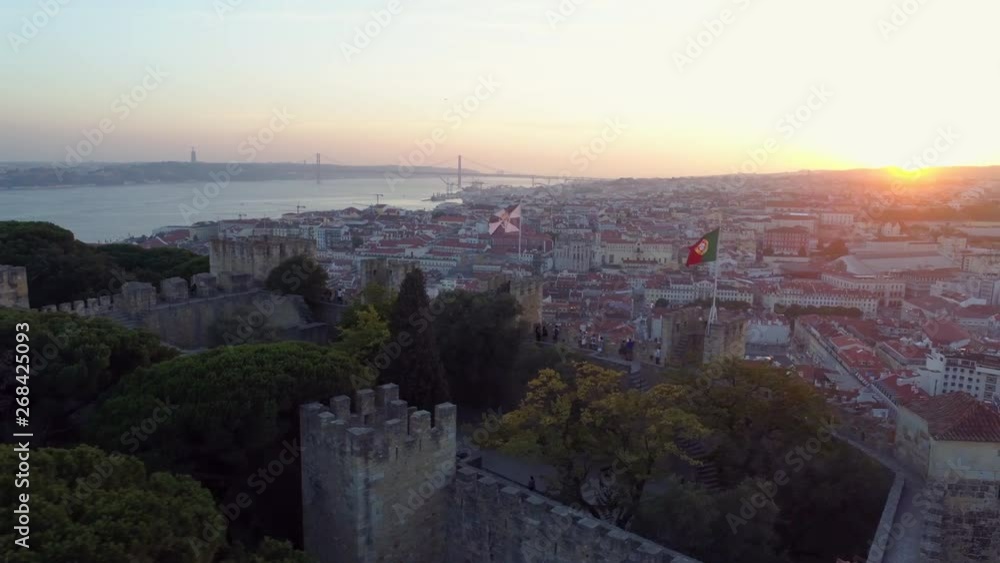 Aerial view medieval fortress on hill top in Lisbon. Drone flying above ancient wall of Castelo de Sao Jorge with flapping flags and walking people at sunset Aerial view medieval fortress on hill top in Lisbon. Drone flying above ancient wall of Castelo de Sao Jorge with flapping flags and walking people at sunset