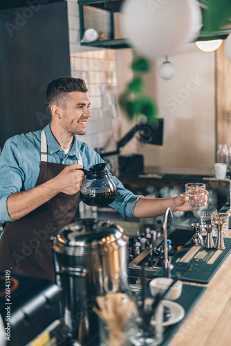 Friendly barista smiling while holding empty glass and coffee jug