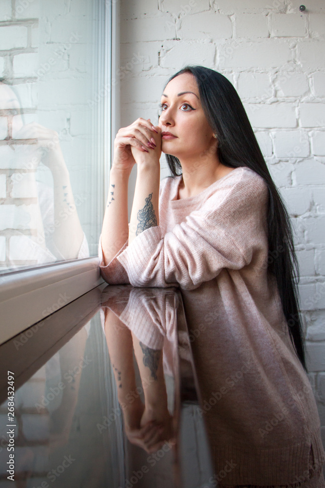 Beautiful young woman with long black hair looks out of the window, reflected in the window and windowsill