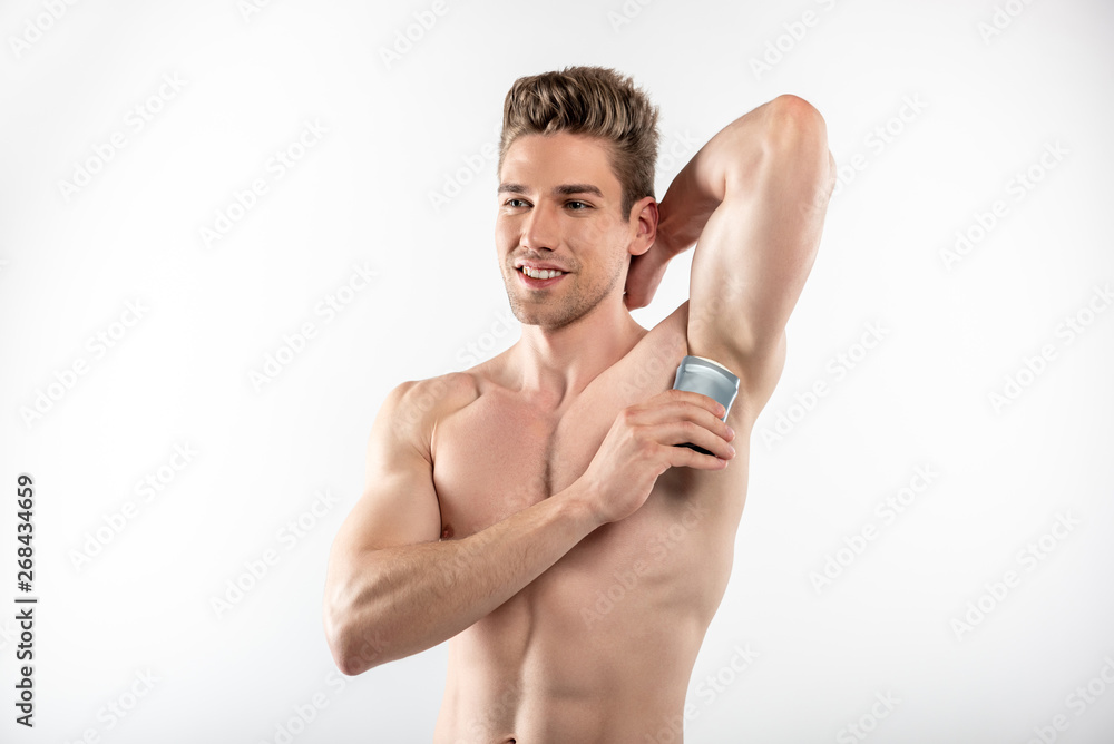 Fototapeta premium Joyful handsome guy with perfect body applying antiperspirant on armpit