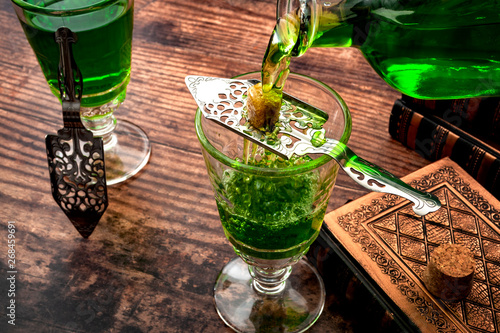 Alcoholic drink, creative stimulant and bohemian lifestyle concept theme with a vintage glass bottle pouring absinthe over a sugar cube in a stainless steel spoon next to books on a wooden table photo
