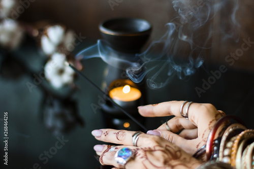 Female wrists painted with henna traditional Indian oriental mehndi ornaments. Hands dressed in metal bracelets and rings holding aromatic stick. Aroma lamp and cotton flowers on background.