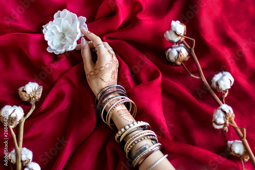 Slender female hand painted with Indian oriental mehndi ornaments by henna. Hand dressed in bracelets hold white flower. Maroon color fabric with folds and cotton branches on background.