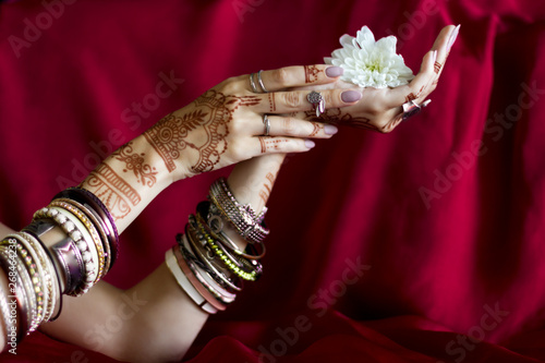 Slender elegant female wrists painted with traditional Indian oriental mehndi ornaments by henna. Hands dressed in bracelets and rings hold white flower. Vinous fabric with folds on background.