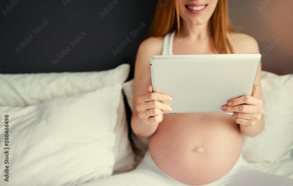 Technology and pregnancy. Young pregnant woman using tablet computer sitting on a bed at home. Technology and pregnancy. Young pregnant woman using tablet computer sitting on a bed at home.