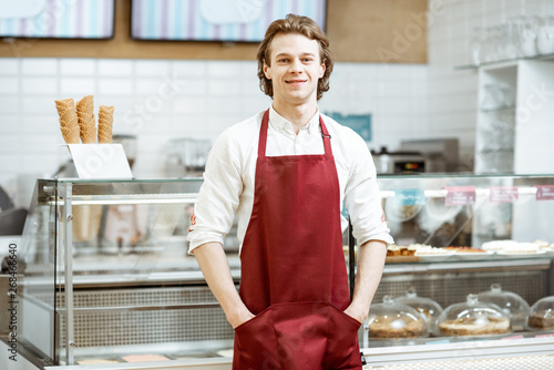 Portrait of a handsome salesman or waiter in red apron standing in the pastry cafe with shop-front on the background photo