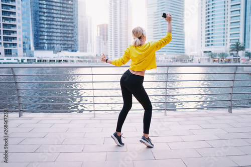 Sport and music. Happy young woman with earphones and smartphone dancing while exercising on city embankment.