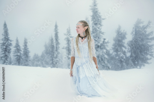 Woman wearing elf ears, dreadlocks and white dress in winter snowy Christmas tree forest. Fog and mystery frozen day