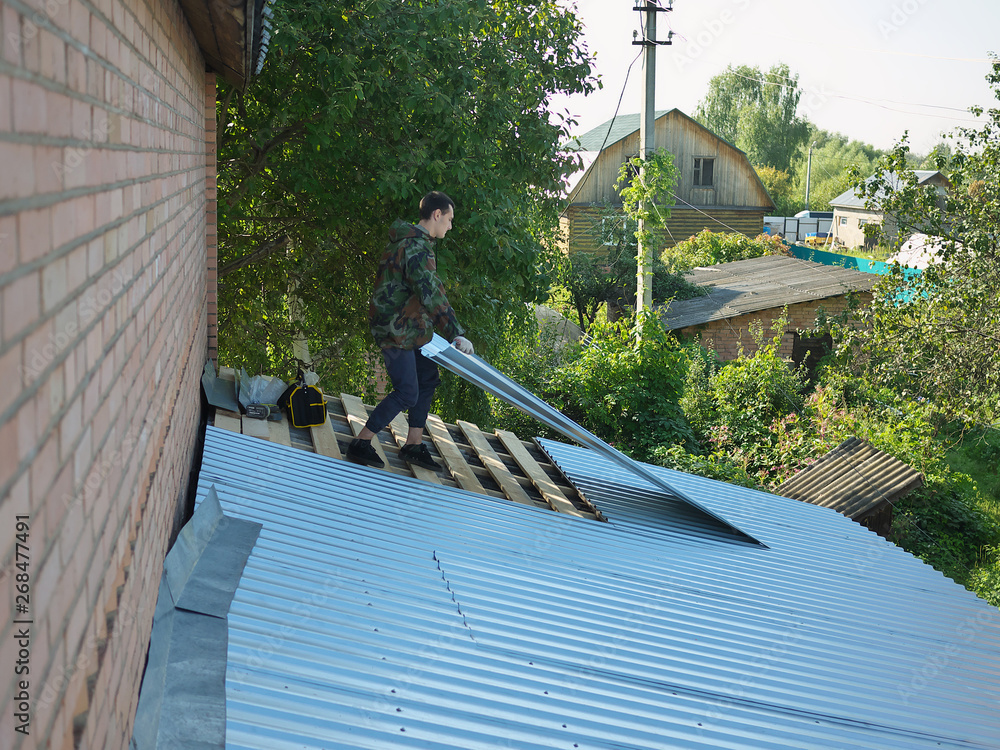 young caucasian man repairing the roof of a home; A worker replaces metal slate on the roof of a home young caucasian man repairing the roof of a home; A worker replaces metal slate on the roof of a home